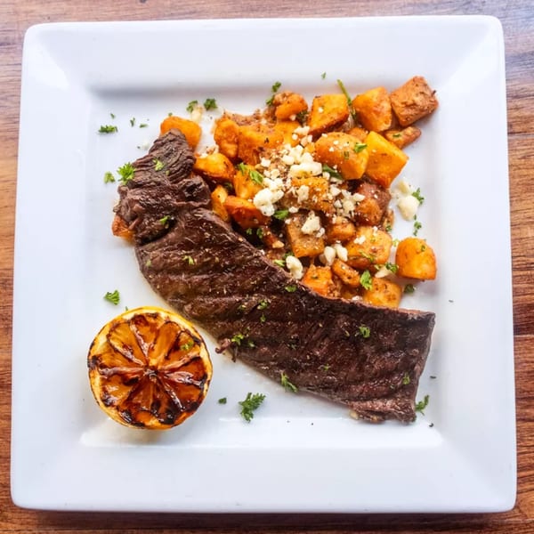 Grilled steak with roasted sweet potatoes, crumbled cheese, and a charred lemon on a white plate.
