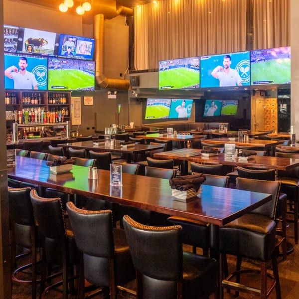A sports bar interior with multiple large screens showing tennis and soccer, high-top tables, and a full bar.