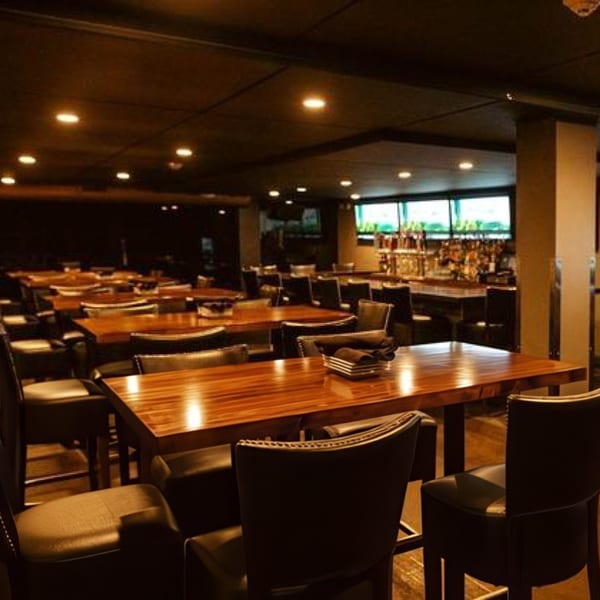 A dimly lit restaurant interior with wooden tables, dark chairs, and a bar in the background.