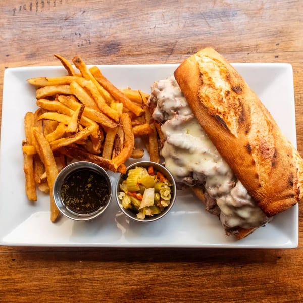 A Philly cheesesteak sandwich with fries, au jus, and giardiniera on a white plate.