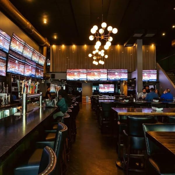 A dimly lit sports bar with multiple large screens, a long bar, and high-top tables.